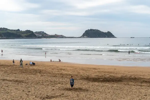Zarautz, İspanya, 4 Nisan 2024: Çocuklar Zarautz 'da kumlu plajda sörfçüler dalgalara binerken ve sakin deniz boyunca yeşil bir ada yükselirken, editoryal görüntü