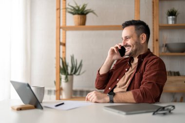 Genç yakışıklı iş adamı modern akıllı telefonu kullanıyor, ev telefonuyla arıyor, masa başında oturuyor, çalışıyor, iletişim kuruyor, müşterilere danışmanlık yapıyor, ticari markalarla daha iyi iletişim kurmalarına yardım ediyor..