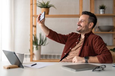 Gülümseyen, serbest çalışan, akıllı telefon kullanan, ön kameraya bakan, gün boyunca ofiste otururken selfie çeken yakışıklı bir erkek..