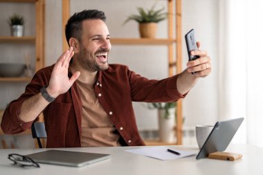 Evden çalışan mutlu Kafkasyalı işadamı, masada oturuyor, ön telefon kamerasına bakıyor, selamlıyor, cep telefonundan birine el sallıyor. Merkez ofis kavramı.