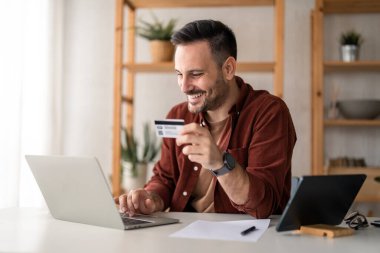 Elinde kredi kartı olan yakışıklı genç bir erkek, gün içinde merkez büroda otururken bilgisayarını kullanarak online bankacılık işlemlerini yapıyor..