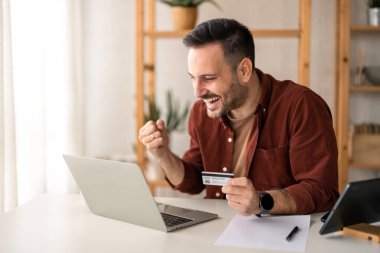 Mutlu genç işadamı, elinde kredi kartı bulunan memnun bir alıcı, dizüstü bilgisayarda online banka uygulaması kullanıyor, uygun finansal e-ticaret ödeme dijital işlemleri yapıyor, evet hareketinde yumruğunu kaldırıyor..