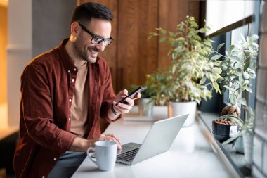Elektronik bankacılık için akıllı telefon ve dizüstü bilgisayar kullanan, internet üzerinden alışveriş yapan ve evde vakit geçirirken ödeme yapan modern çağdan memnun bir adam..