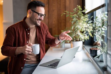 Neşeli, yakışıklı, kendine güveni tam bir adam uzaktan kumandalı online iş görüşmesine ya da iş görüşmesine gidiyor. Elinde bir fincan kahveyle evden gelen sanal video konferansında el kol hareketi yapıyor..