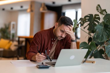 Contemplating businessman in the home office feeling sad. Full concentration on work. Man working on laptop while sitting at his working place in his apartment. Tired brown haired man taking glasses off working too long at computer. Exhausted male su clipart