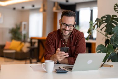 Mutlu güler yüzlü iş adamı günlük giysiler giyer ve sabah erken saatlerde evinde modern akıllı telefon kullanır, başarılı bir işveren iş yerinde otururken anlaşma yapar..