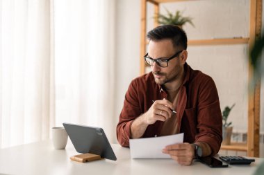 Genç ciddi meşgul profesyonel işadamı ya da öğrenci dijital tablet kullanarak gözlük takıyor online webinar izliyor ya da web eğitimi alıyor, tablete bakıyor, düşünüyor, yazıyor, araştırma yapıyor..