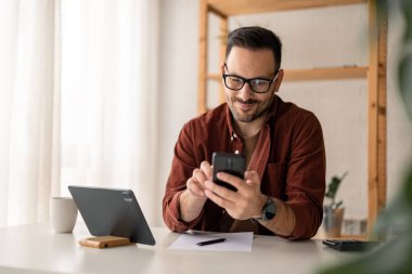 Mutlu gülümseyen iş adamı günlük giysiler giyer ve gün içinde evinde modern akıllı telefon kullanır, daktilo eder, ekrana dokunur, internette dolaşır ya da mesaj yazar..