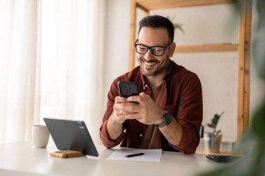 Mutlu gülümseyen iş adamı günlük giysiler giyer ve ev ofisinde modern akıllı telefon kullanır, başarılı işveren masasında oturur cep telefonuyla yazar, ayrıca dijital tablet, kalem ve kağıt kullanır..