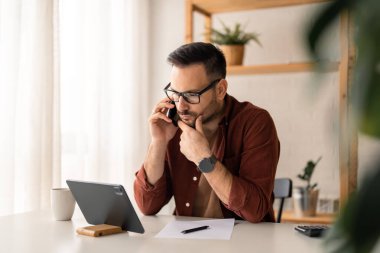 Yakışıklı genç işadamı, borsacı ya da danışman dahili ofisten akıllı telefondan konuşuyor, müşteriye danışıyor, dijital tablete bakıyor finansal veri yatırımı hakkında düşünüyor..