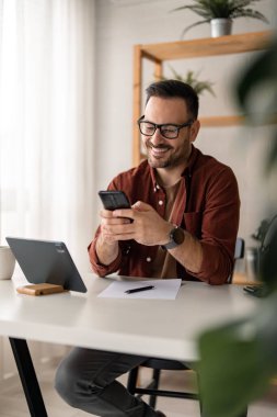 Mutlu gülümseyen iş adamı günlük giysiler giyer ve ev ofisinde modern akıllı telefon kullanır, başarılı işveren masasında oturur cep telefonuyla yazar, ayrıca dijital tablet, kalem ve kağıt kullanır..