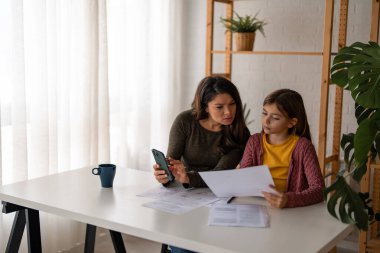 Genç kahverengi saçlı anne ve ödemeleri kontrol eden kızı. Ev ekonomisi konsepti. Oturmak ve ödemeleri evden yapmak..