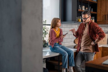 Angry strict father scolding stubborn daughter for spending too much time online on a mobile phone. Father and daughter having a discussion at home. Family conflicts and punishment concept clipart