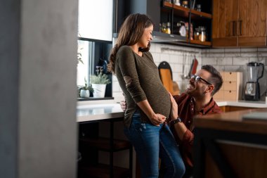 Gözlüklü, gülümseyen bir adamın, evde bir mutfakta hamile karısının karnına nazikçe dokunuşunun yan görüntüsü..