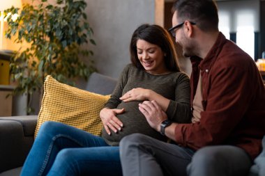 Koca karısına sarılıyor ve hamile karnında el ele tutuşuyor. Birbirlerine bağlanıyorlar, evdeki kanepede oturuyorlar..
