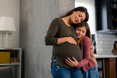 Mutlu hamile anne, sevimli genç kızıyla eğleniyor. Evde durmuş, gözleri kapalı gülümserken kucaklaşıyor. Hamilelik boyunca aile sevgisi ve destek.