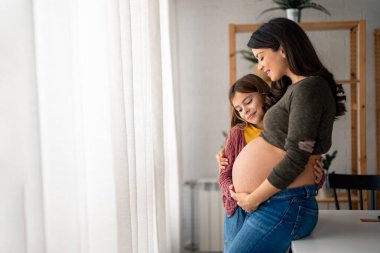 Çok tatlı bir kız annesinin hamile karnına dokunuyor. Mutlu genç kız, annesinin karnında bebeği hissederken onu evinde kucaklıyor. Güzel hamile anne güzel kızıyla pencerenin yanında duruyor..