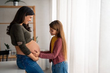 Kıvırcık saçlı, genç, hamile bir kadın. Sevgi dolu gözleriyle sevimli kızına bakıyor ve yüzüne gülüyor. Tatlı küçük kız evde hamile bir anneyle bağ kuruyor, bebeği hayal ediyor..