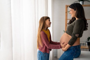Evinde genç bir ilkokul kızı olan hamile ve gülümseyen bekar bir anne. Uzun saçlı tatlı kız nazikçe annemin karnına dokunuyor. Hamile kadın ve küçük kız birbirlerine bakıyorlar..