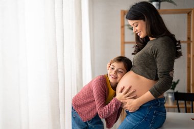 Güzel, hamile, bekar bir anne hamile karnına yaslanmış tatlı küçük kızını nazikçe kucaklıyor. Meraklı güzel kız, annesinin karnında bebeği hissetmeye çalışıyor..