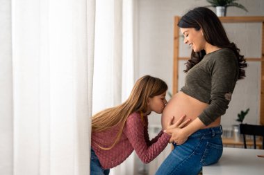 Hamile bir annenin göbeğini öpen sevimli küçük kız. Hamileliğin ileri safhasında, evde neşeyle kaynaşan güzel, bekar bir anne. Önemseyen küçük kız annesinin karnında bebeğe öpücük veriyor..