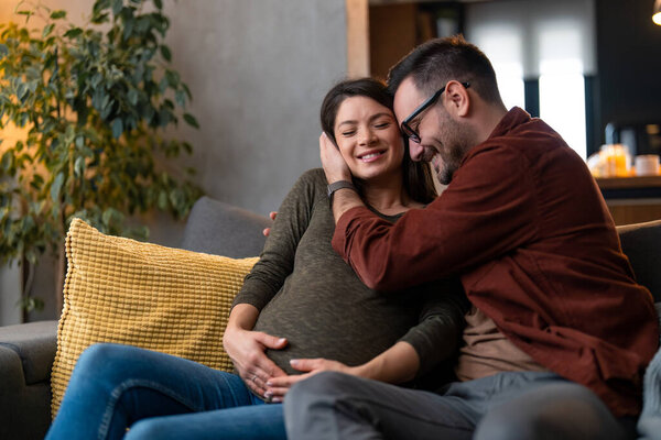 Supportive and caring husband gently hugging his pregnant wife at home while spending time together at home, sitting on a sofa, enjoying in peaceful moments.
