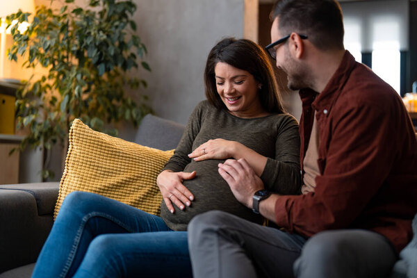Муж обнимает жену и держит за руки ее беременный живот. Они сближаются, сидят дома на диване.