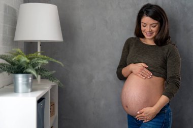 Koyu yeşil bluz ve kot pantolon giyen güzel müstakbel anne yüzünde bir gülümsemeyle nazikçe karnına bakıyor ve ona dokunuyor..