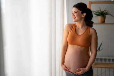 Hamileliğinin son üç ayında genç ve güzel bir kadın yoga pantolonu giyiyor ve turuncu spor sutyeniyle evinin penceresinden bakarken el ele tutuşuyor..