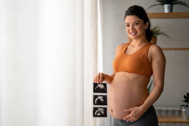 Göbeğini gösteren rahat giysiler içinde güzel bir gülümsemesi olan mutlu hamile bir kadın. Bir elinde ultrason resimleri, diğer elinde ise karnının üstünde..