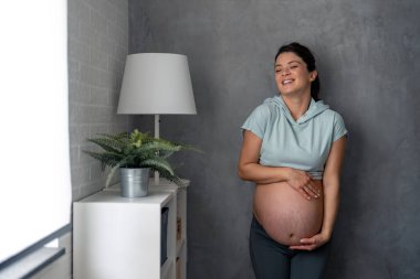 Yumuşak mavi tişört ve gri tayt giyen neşeli hamile kadın bebeği hayal ediyor, karnını tutuyor ve gülümsüyor..