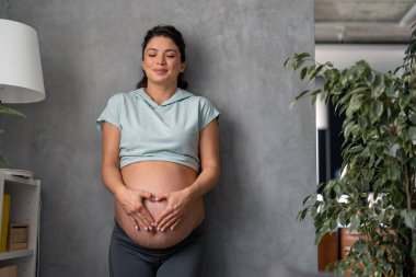 Hayalperest anne yumuşak mavi tişört ve gözleri kapalı gri taytlar giyer ellerini karnına koyar ve kalp şekli oluşturur..