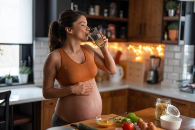 Hamile kadın mutfakta durmuş kahvaltı hazırlarken bir bardak su içiyor..