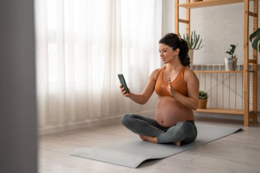 Evde yoga yaparken telefonu kullanan hamile bir kadın.