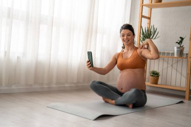Neşeli, mutlu, hamile bir kadın evde yoga minderinde egzersiz yaparken akıllı telefon uygulaması kullanıyor..