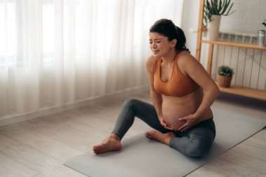 Mutsuz Kafkasyalı, evinde egzersiz minderinde otururken karın ağrısı çeken bir kadın bekliyor..