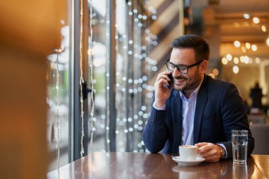 Profesyonel kıyafetli, orta yaşlı, neşeli bir beyaz adam elinde kahve fincanıyla, iyi aydınlatılmış bir kafe masasında otururken akıllı telefonuyla konuşuyor..