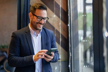 Gülümseyen orta yaşlı, beyaz, şık giyimli bir işletme yöneticisi akıllı telefonunu çağdaş bir kafe içinde ustalıkla gezdiriyor..