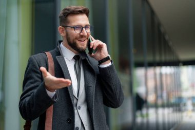 Sakallı, neşeli, orta yaşlı bir işadamı, iş kıyafeti giymiş, akıllı telefonuyla konuşurken aktif bir şekilde el kol hareketi yapan bir telefon alıyor..
