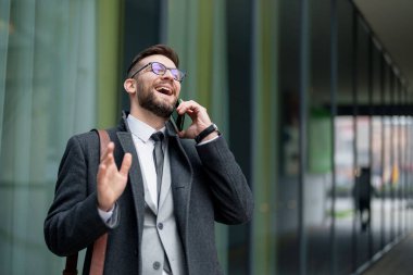 Zeki giyimli neşeli, orta yaşlı bir işadamı, modern bir binanın yanında duran akıllı telefonuyla, yoğun bir profesyonel yaşam tarzını yansıtan bir çağrı yapıyor..