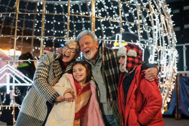 Gülümseyen yaşlı bir çift, parıldayan ışıklarla çevrili açık hava ortamında genç torunlarını kucaklıyor ve bir kutlama sırasında ailenin sıcaklığını işaret ediyorlar..