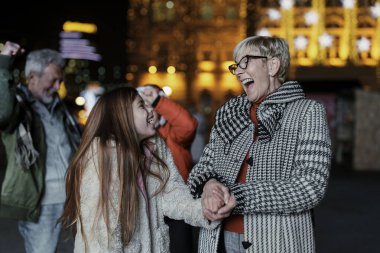 A joyful Caucasian grandmother laughs with her granddaughter, both bundled in warm attire, as they share a festive moment surrounded by illuminated decorations outdoors in an urban environment. clipart