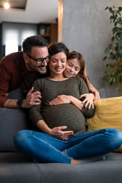 Маленькая девочка и заботливый муж обнимают мать с любовью и нежностью сзади, пока она сидит на диване