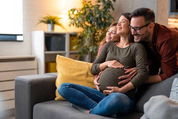 Smiling pregnant mother enjoying the emotional support during pregnancy from her husband and daughter. Happy healthy family embracing at home, excited about the new family member.