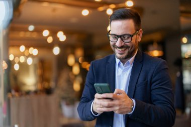 İş kıyafetleri içinde neşeli, orta yaşlı bir beyaz adam akıllı telefonuna odaklanırken modern bir kafenin davetkar atmosferinin tadını çıkarıyor..