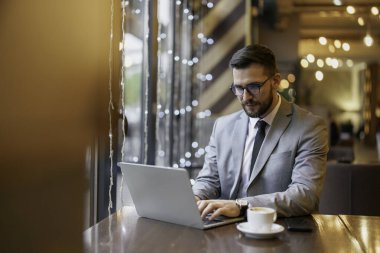 Sivri takım elbiseli, düşünceli, beyaz bir erkek yönetici, iyi aydınlatılmış çağdaş bir kafe ortamında, dizüstü bilgisayarıyla yan yana duruyor..