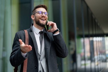 Sırt çantalı şık bir takım elbise giymiş, gülümseyen beyaz bir işadamı, modern bir binanın dış cephesi tarafından cep telefonuyla canlı bir sohbete girişir..