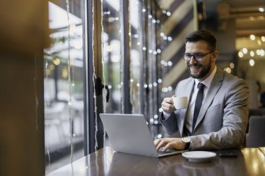 İş kıyafeti giymiş orta yaşlı beyaz bir erkek modern bir kafe ortamında kahvenin tadını çıkarırken dizüstü bilgisayarda çalışıyor..