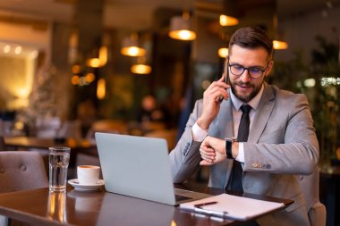 Takım elbiseli yakışıklı iş adamı akıllı telefondan konuşuyor, el saatine bakıyor, saati kontrol ediyor, modern restoranda oturuyor iş ortağının iş yemeğine gelmesini bekliyor..