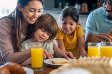 Gülümseyen genç anne çocuklarına kahvaltı hazırlarken onlarla ve kocasıyla birlikte masada oturuyor..
